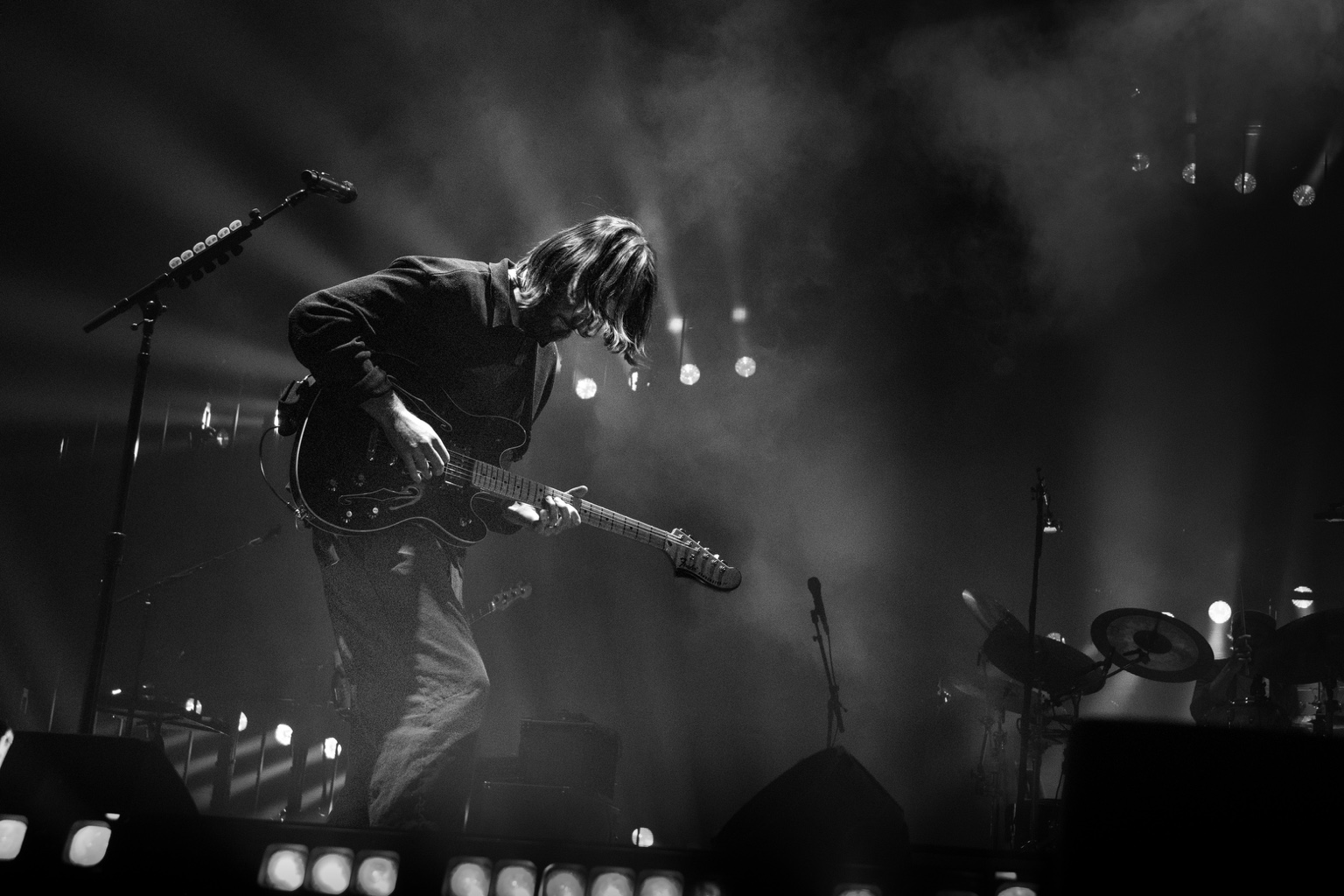 A guitarist leans into his instrument beneath beams of light on a smoky stage
